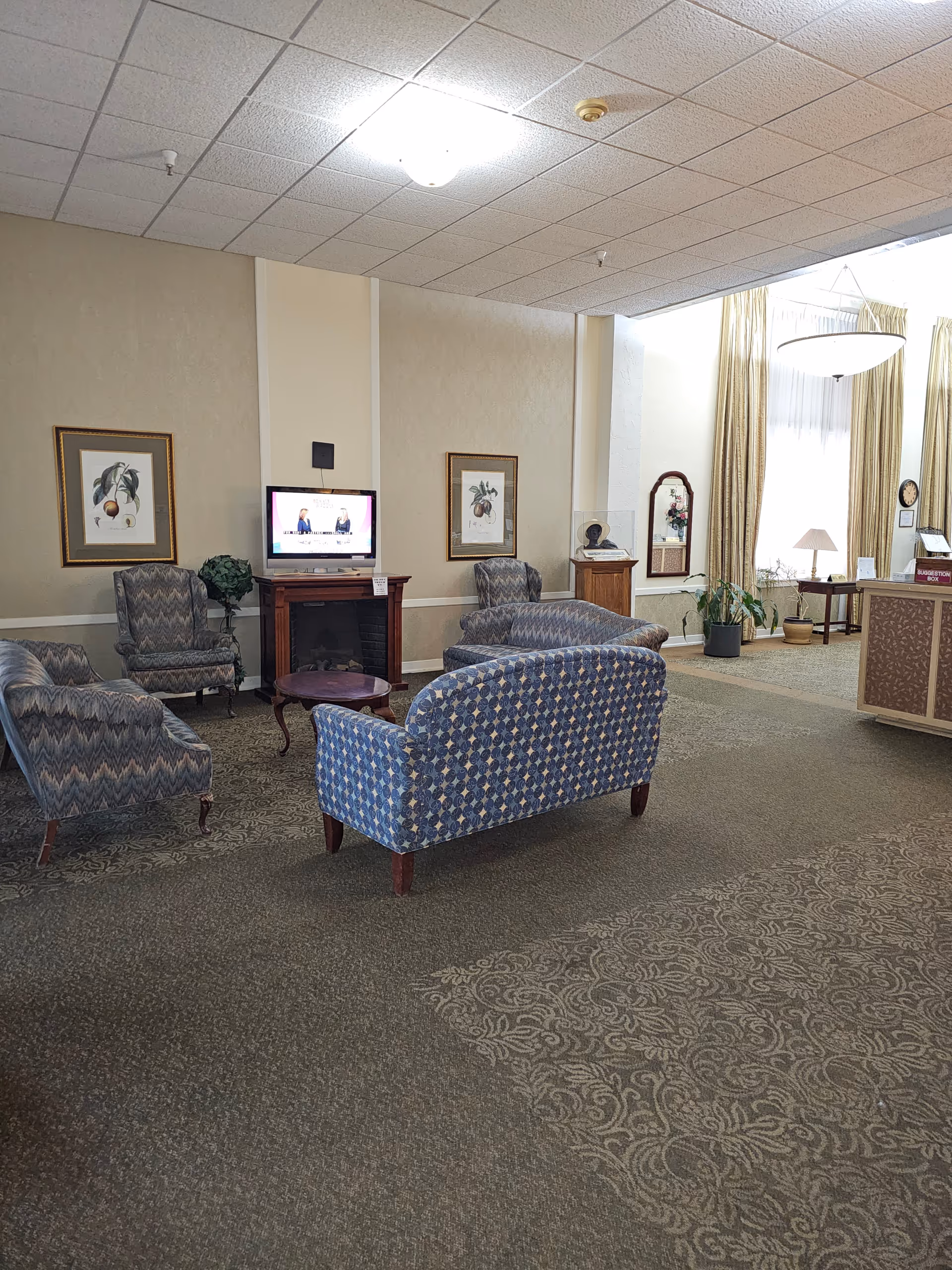 A senior living facility common area with patterned upholstered chairs and sofas arranged around a small wooden table. A flat-screen TV is mounted on the wall above a wooden fireplace. The room has beige walls, framed botanical prints, a carpeted floor with a floral pattern, and large windows with long curtains letting in natural light. There is a reception desk to the right with a suggestion box and a lamp on a side table near the windows.
