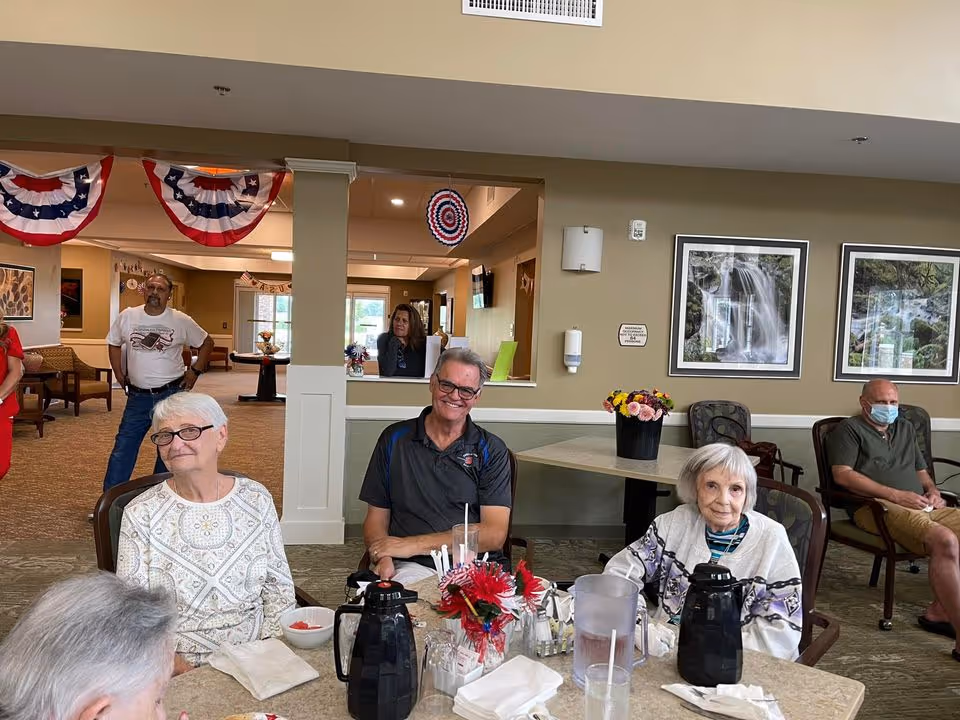 A group of elderly people sitting around a table in a common area of an assisted living facility. The table has coffee pots, glasses, napkins, and a small flower arrangement. In the background, there are patriotic decorations hanging from the ceiling, framed pictures on the wall, and a few other people standing or sitting. One man is wearing a face mask.