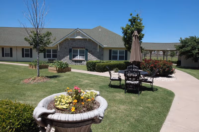 Sunlit courtyard with a flower-filled planter, a patio table and umbrella on a lawn in front of a single-story assisted living building.