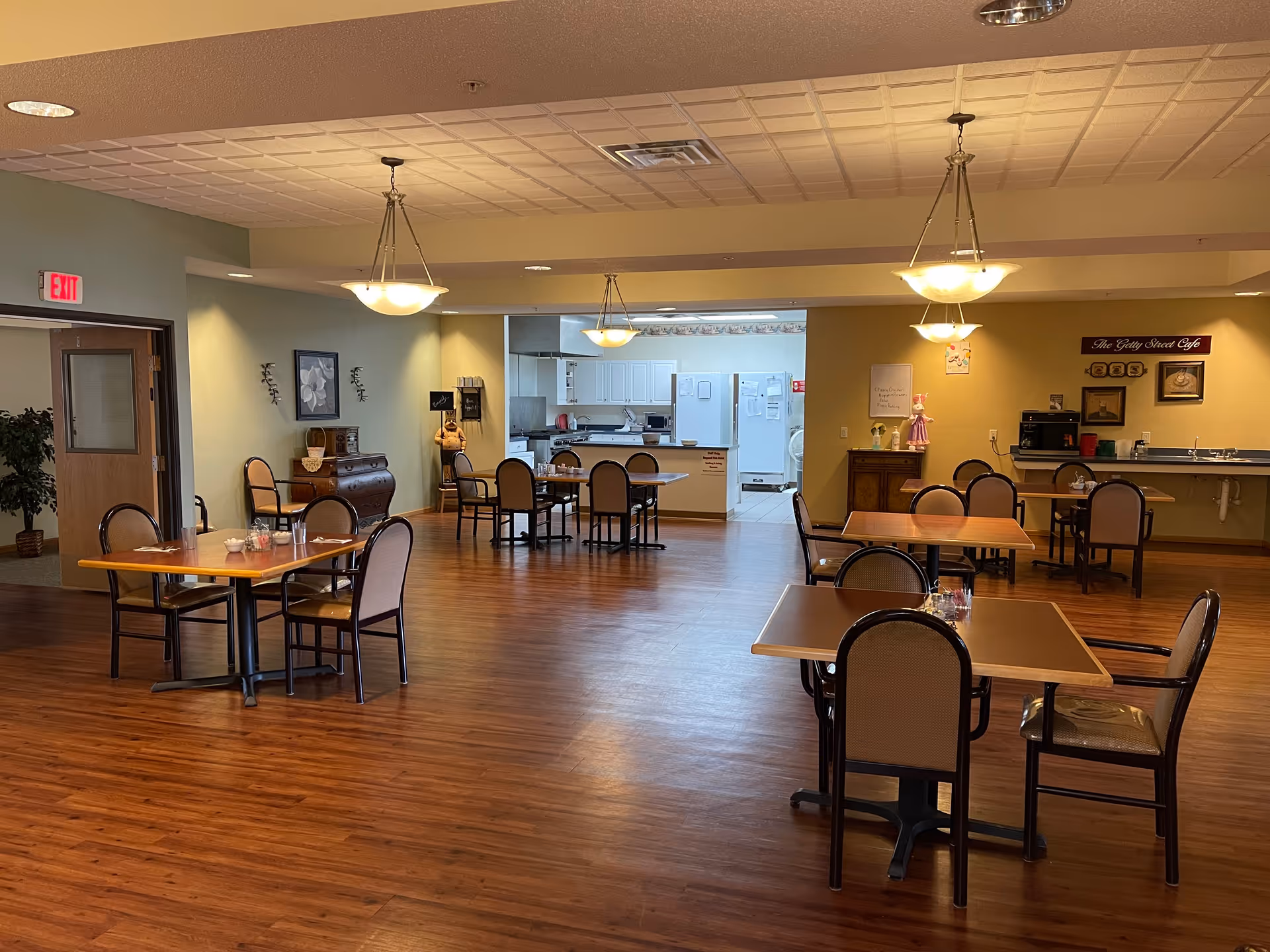 A dining area in an assisted living facility with several tables and chairs arranged on a wooden floor. The room is well-lit with hanging ceiling lights. In the background, there is an open kitchen area with white cabinets and appliances. The walls are painted in soft green and yellow tones, and there are decorative items and framed pictures on the walls. A sign on the yellow wall reads 'The Getty Street Cafe'.