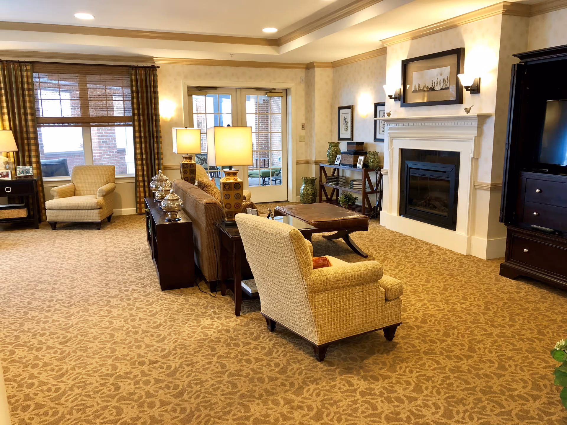 Bright, carpeted communal living room with armchairs and sofas arranged around a coffee table and fireplace, with lamps and large windows.