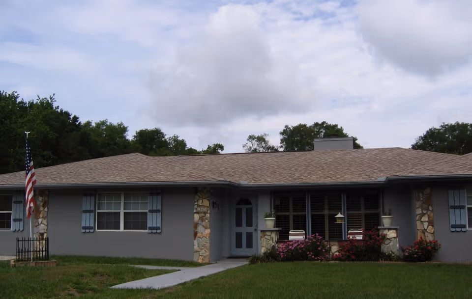 Single-story ranch-style house with a central front door, shuttered windows, stone accents, porch planters and an American flag on the lawn.