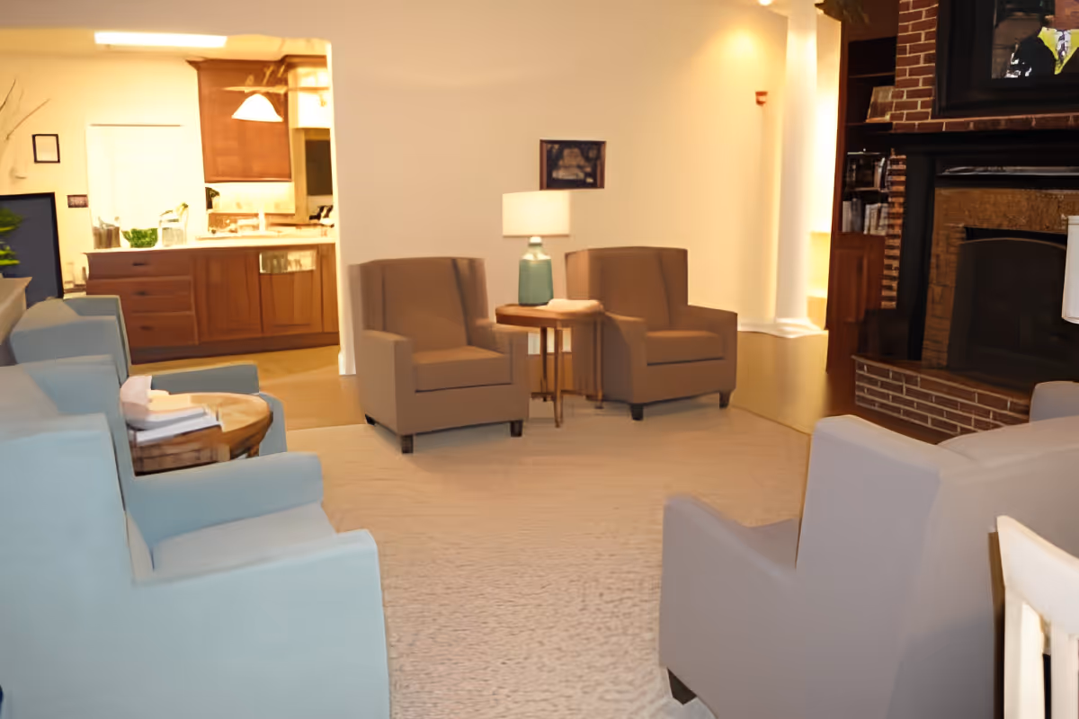 A cozy living room area with several armchairs arranged around a light-colored carpet. There is a small wooden side table with a lamp between two beige armchairs. In the background, a kitchen area with wooden cabinets and a countertop is visible. A brick fireplace with a TV mounted above it is on the right side of the room.