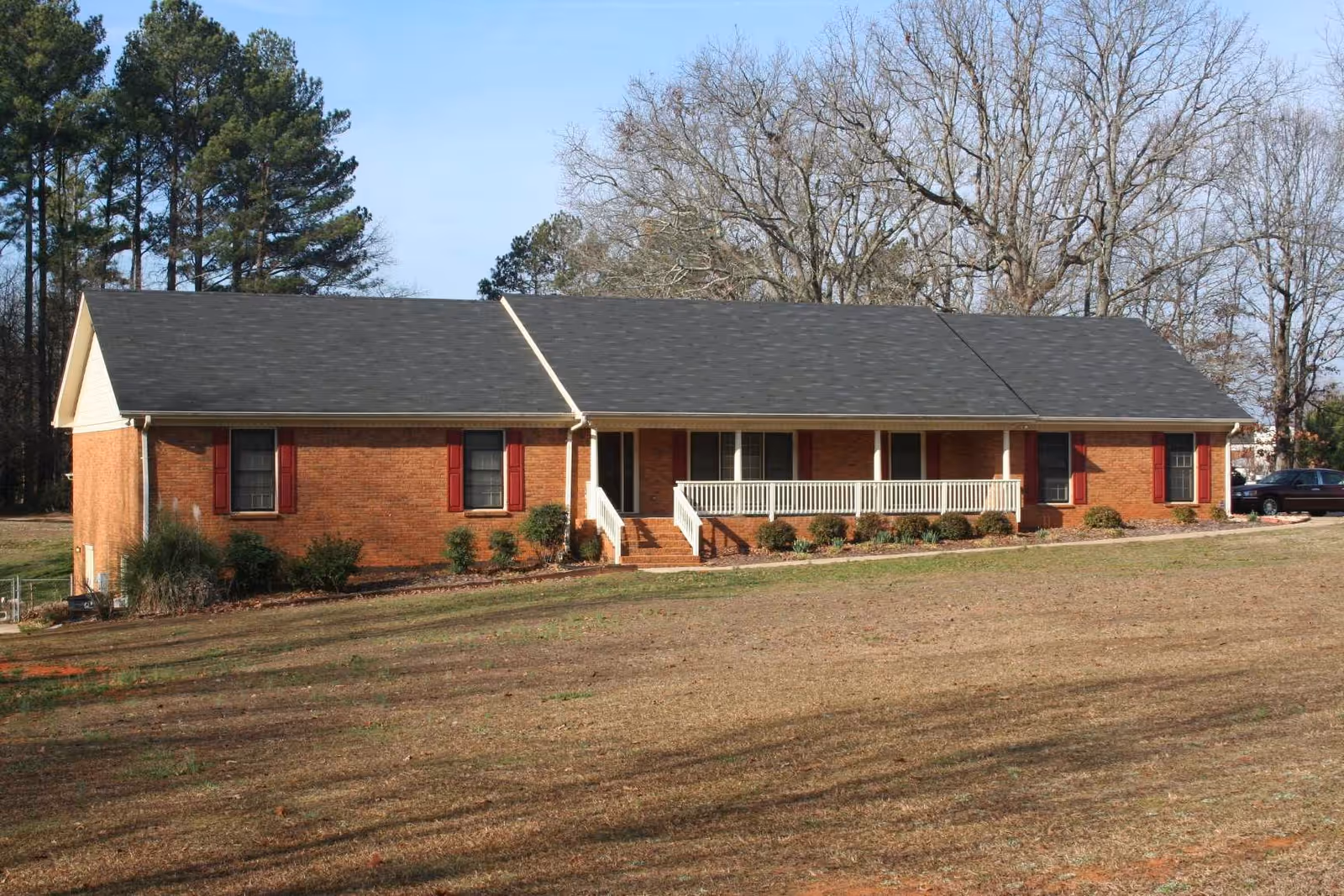 Single-story brick building with a dark shingled roof, white porch railing, and red window shutters, surrounded by a grassy yard and trees in the background.
