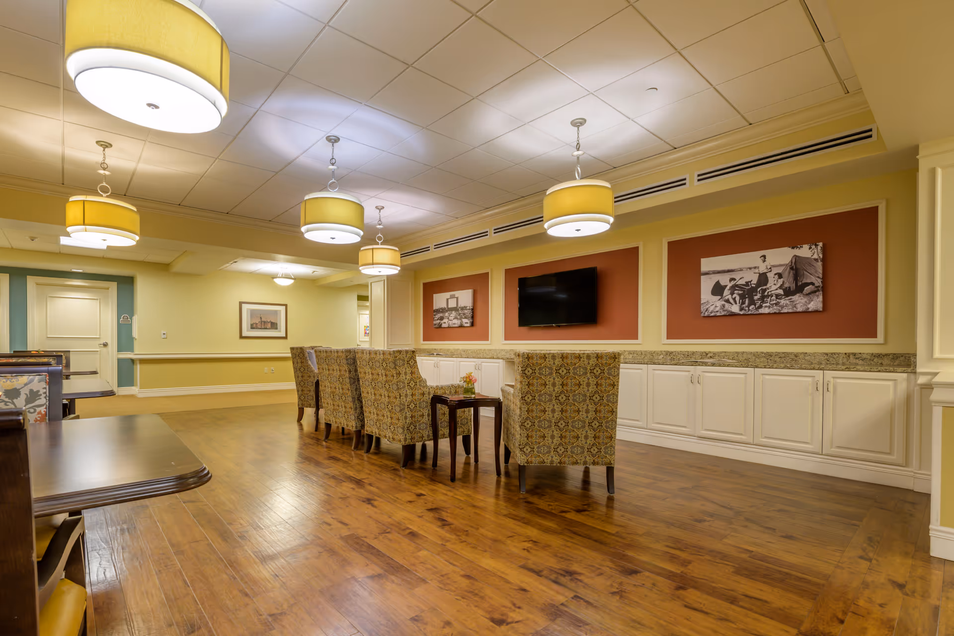 A spacious, well-lit common area with wooden flooring, patterned armchairs arranged around a small table, a wall-mounted flat-screen TV, and framed black-and-white photographs on the walls. The ceiling features multiple round pendant lights with yellow shades.