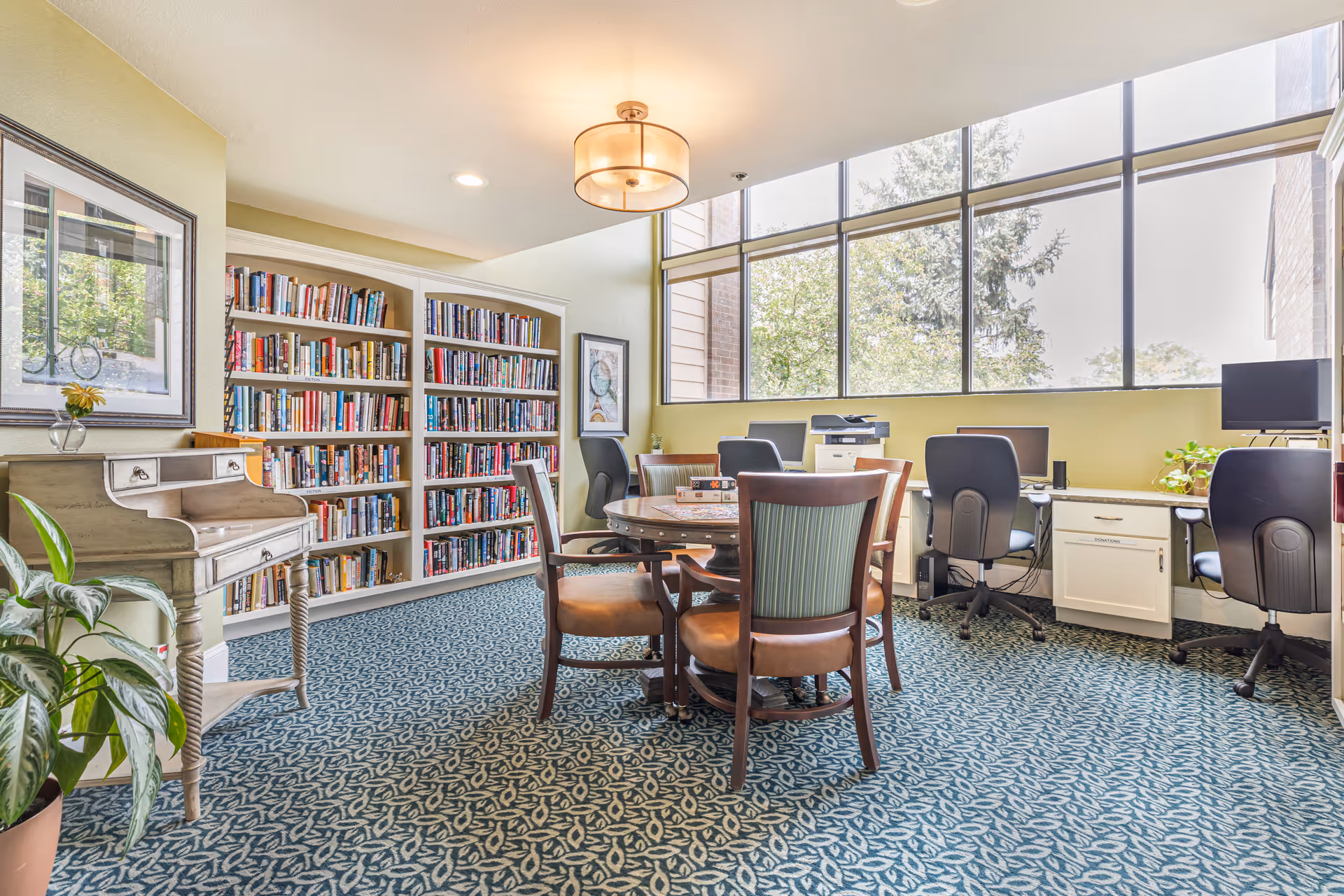 A bright room with large windows showing trees outside. Inside, there is a round wooden table with four chairs around it, a bookshelf filled with books, a small vintage-style desk with drawers, and a row of computer workstations with office chairs. The room has patterned blue carpet and light green walls.