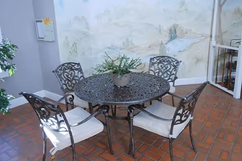 A round metal table with intricate designs surrounded by four matching metal chairs with white cushions, placed on a brick floor. A potted plant is centered on the table. The background features a wall with a soft mural of a natural landscape.