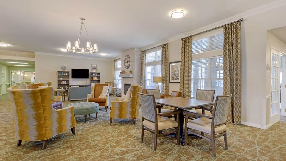A spacious and well-lit senior living common area with patterned carpet, several upholstered armchairs, a round ottoman, a rectangular wooden table with six chairs, large windows with patterned curtains, a fireplace with a clock above it, and a TV mounted on the wall with bookshelves on either side.