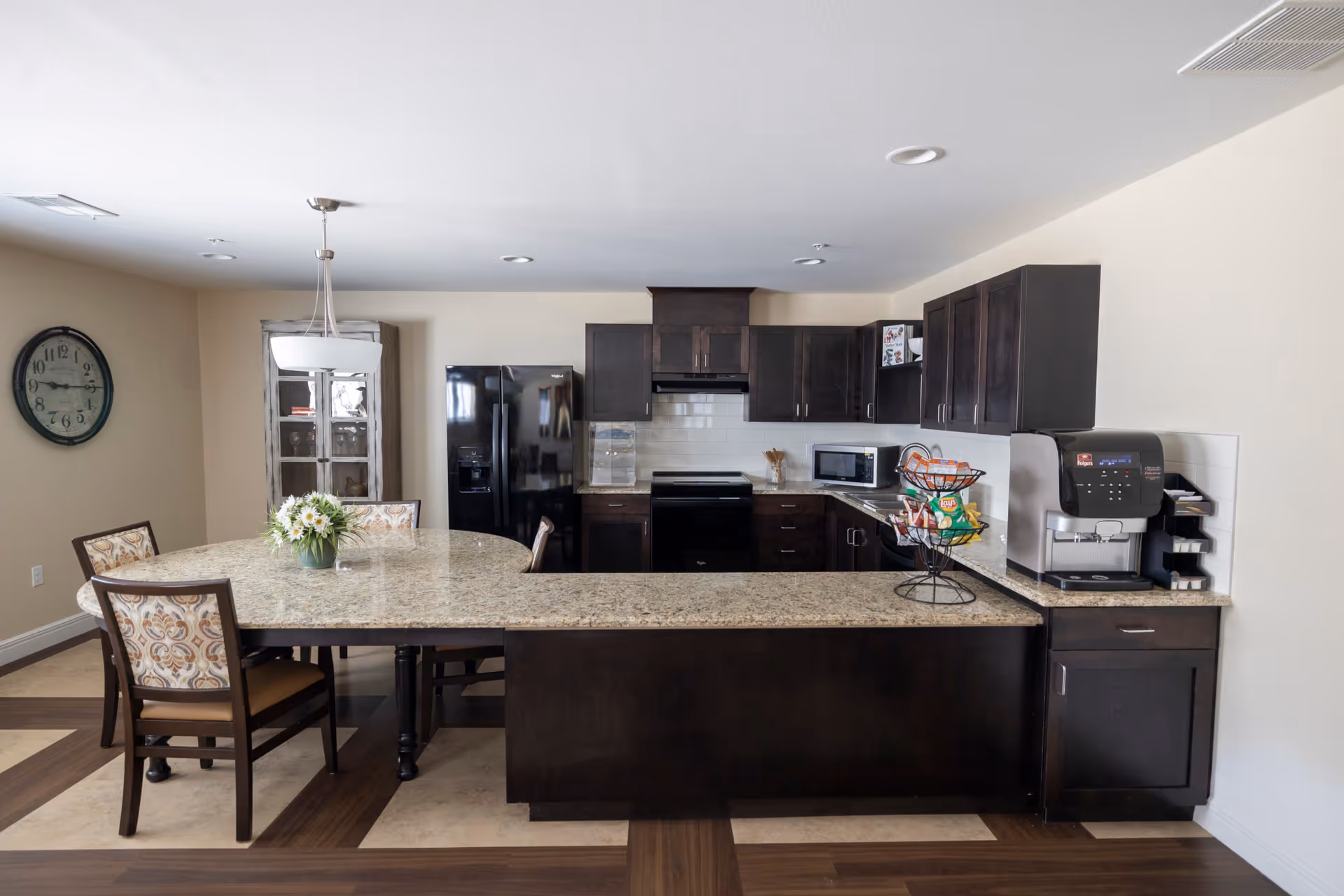 A modern kitchen area in a senior living facility featuring dark wood cabinets, a black refrigerator, stove, and microwave. There is a large granite countertop island with a vase of flowers and a tiered snack holder. Four chairs with patterned cushions surround a dining table attached to the island. A wall clock and a hanging light fixture are visible on the beige walls.