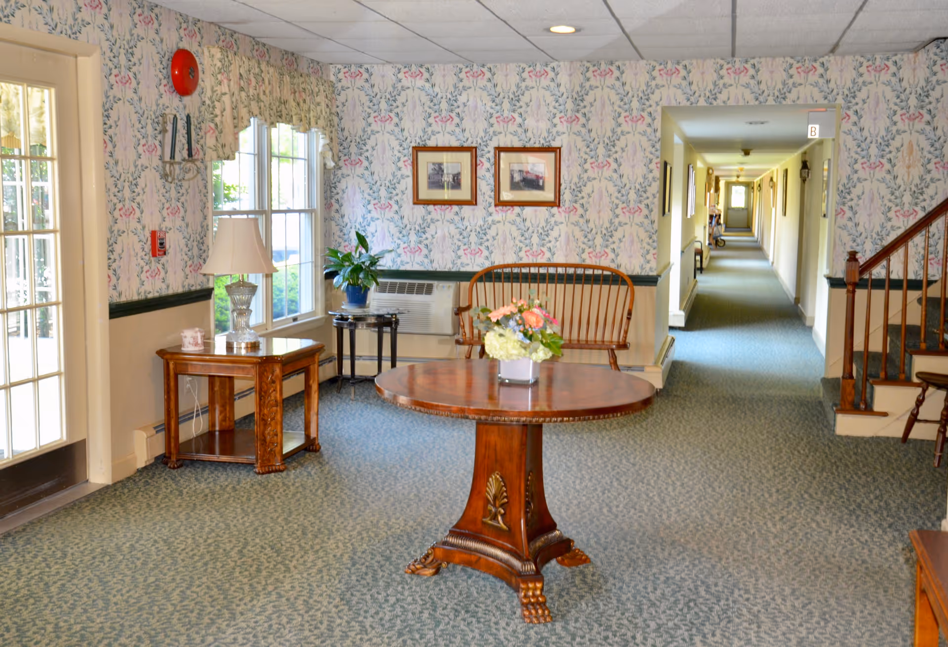 A cozy interior common area with floral wallpaper, a round wooden table with a flower arrangement, a wooden bench, side tables with a lamp and a plant, and a long hallway leading to other rooms. There is a staircase on the right side and large windows letting in natural light.