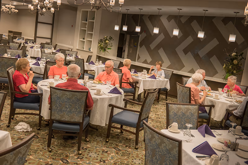 A dining room in a senior living facility with several elderly people seated at round tables covered with white tablecloths. The tables are set with plates, glasses, and purple folded napkins. The room has patterned carpet, decorative wall art, and hanging pendant lights.