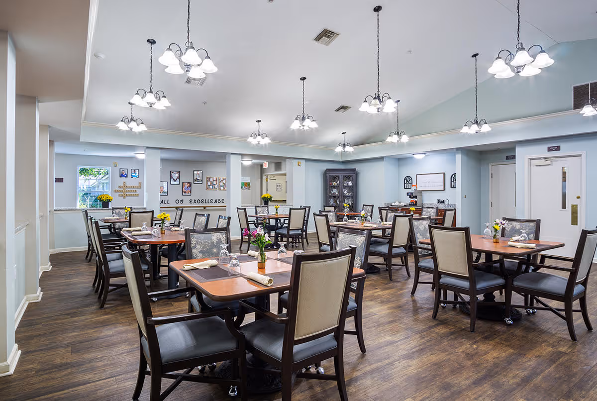 A spacious dining room with multiple wooden tables and cushioned chairs arranged neatly. Each table is set with placemats, napkins, glassware, and small flower vases. The room has wooden flooring, light blue walls, and several hanging light fixtures providing bright illumination. There are decorative elements on the walls and a cabinet in the corner.