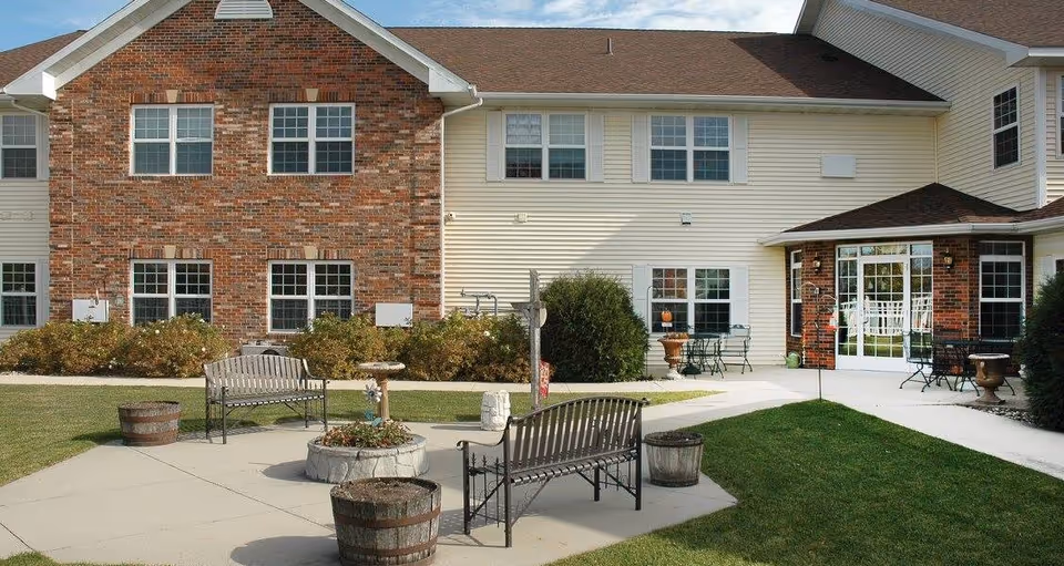 Outdoor patio area at Oak Park Place Albert Lea featuring two metal benches, a small round planter with flowers, several wooden barrel planters, and a birdbath. The patio is surrounded by a well-maintained lawn and shrubbery, with a two-story building in the background that has a combination of brick and light-colored siding, multiple windows, and glass doors leading to the patio.