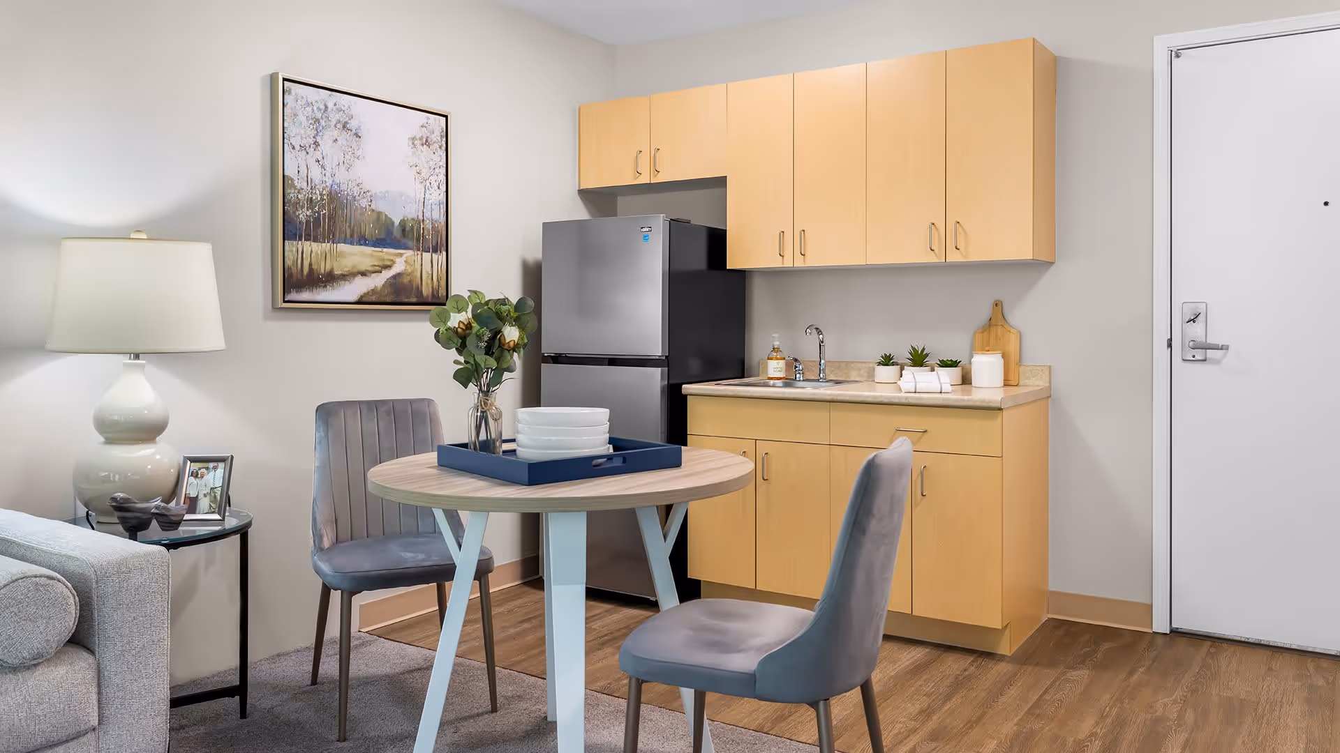 A small kitchenette area with light wood cabinets, a stainless steel refrigerator, and a countertop with a sink. In front of the kitchenette is a round wooden table with two gray upholstered chairs. On the table is a blue tray holding white bowls and a vase with green foliage. To the left is a gray sofa with a side table holding a lamp and a framed photo. A landscape painting hangs on the wall above one of the chairs. The floor is wood with a small carpet under the table, and a white door is visible on the right side.