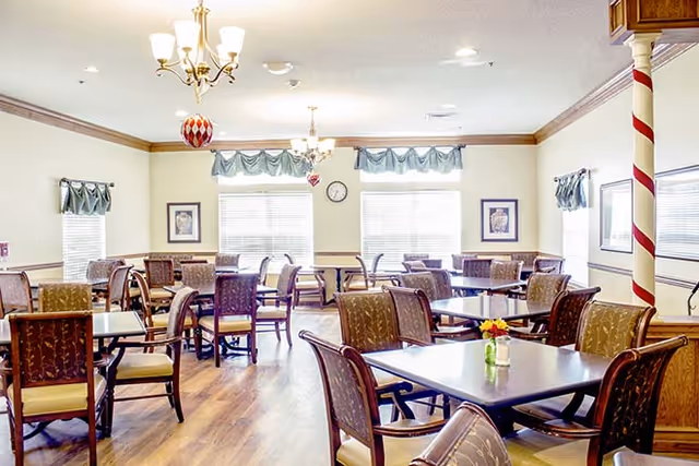 A bright dining room with multiple tables and chairs arranged neatly. The room has large windows with valance curtains, framed artwork on the walls, and two chandeliers hanging from the ceiling. A candy cane striped pillar is visible on the right side, and a small flower vase is placed on one of the tables.
