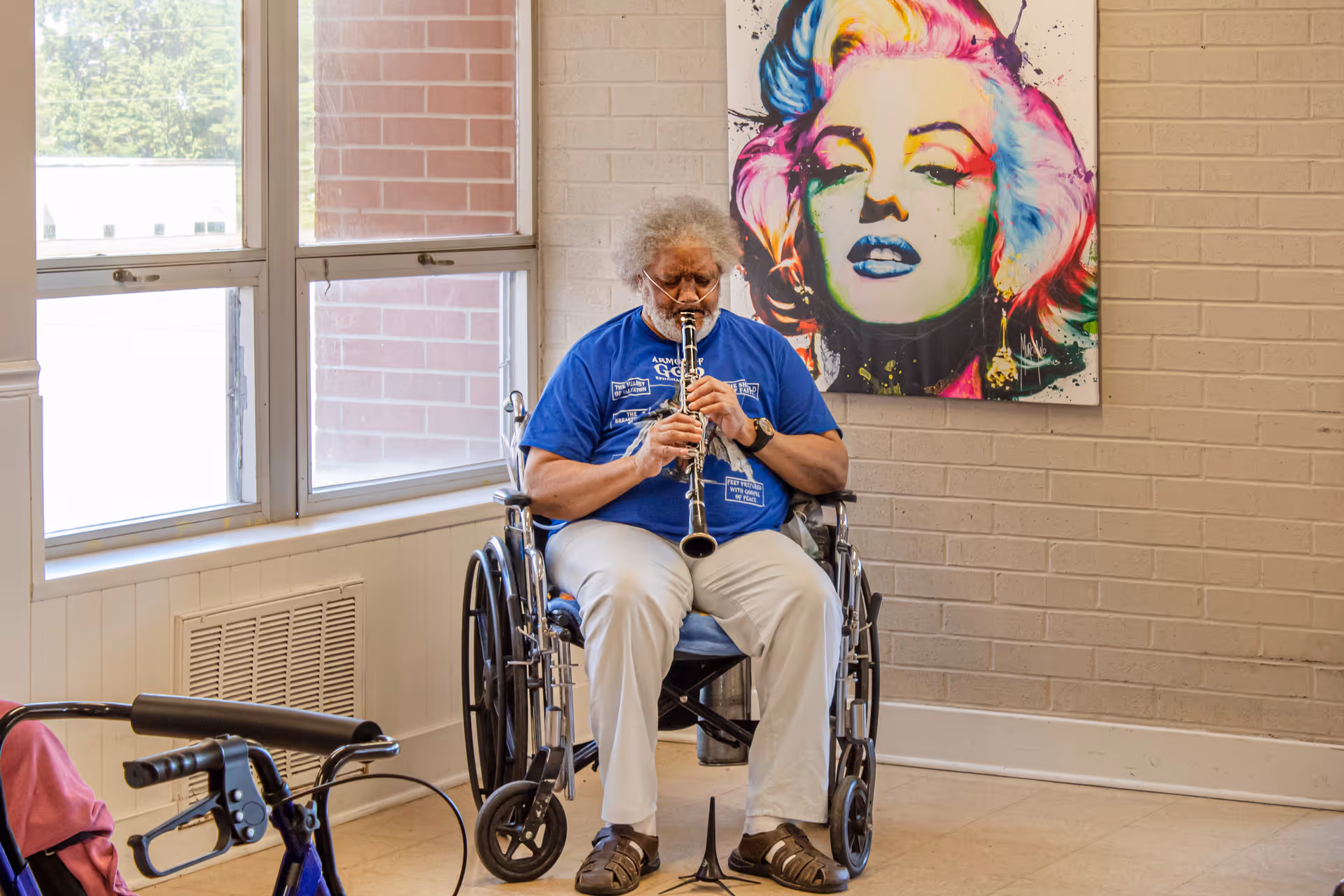 An elderly person sitting in a wheelchair playing a clarinet in a room with large windows and a colorful pop art painting of Marilyn Monroe on the wall behind them.