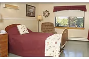 A small assisted-living bedroom with a single adjustable bed covered in burgundy bedding, a nightstand, upholstered chair, lamp, window with a valance, and wall decor.