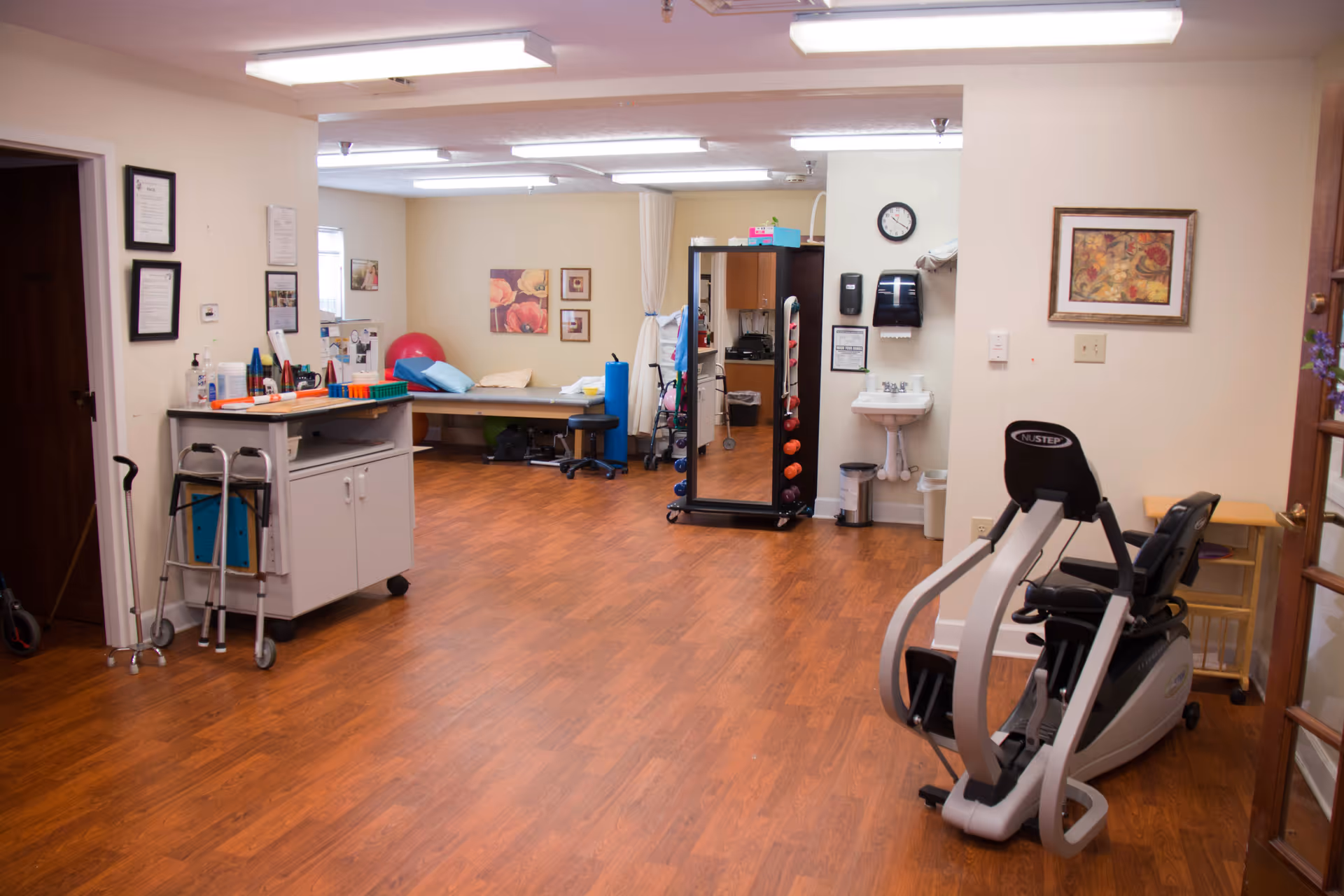 A physical therapy or rehabilitation room with wooden flooring, exercise equipment including a NuStep machine, a walker, a cane, a treatment table with pillows, exercise balls, a mirror with dumbbells, a small sink with soap and paper towel dispensers, and framed artwork on the walls.