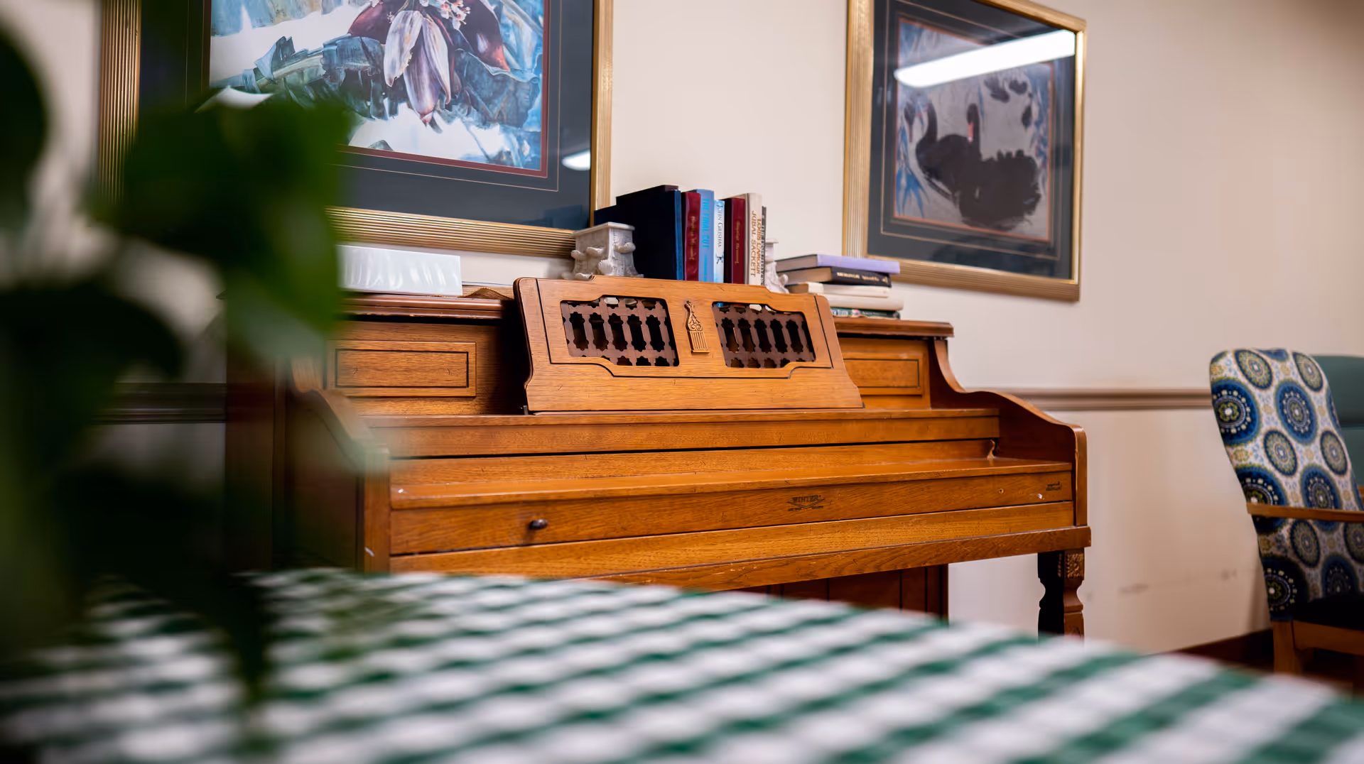 A wooden piano with books and decorative items on top, situated against a wall with framed artwork. A patterned chair is partially visible to the right, and a green and white checkered tablecloth is in the foreground.