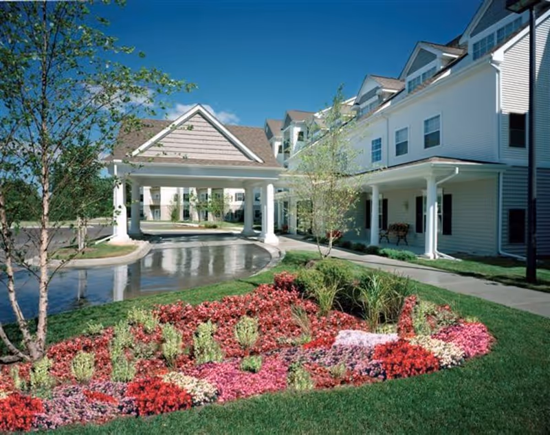 Front entrance of a multi-story senior living building with a covered porte-cochère, manicured flowerbeds and lawn.