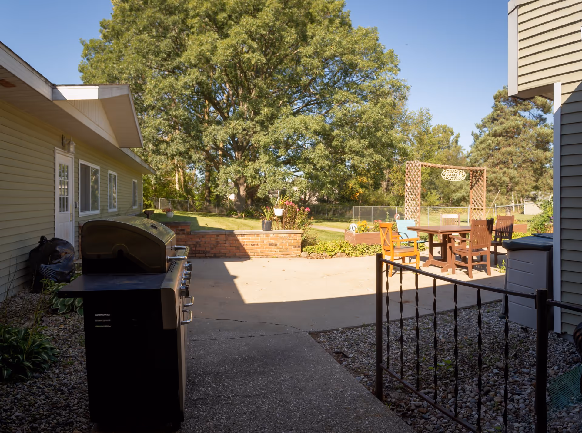 Outdoor patio area at Americana Seniors featuring a large barbecue grill on the left, a concrete patio with a wooden table and chairs, raised garden beds with flowers, a decorative wooden arch with a welcome sign, and large green trees in the background under a clear blue sky.
