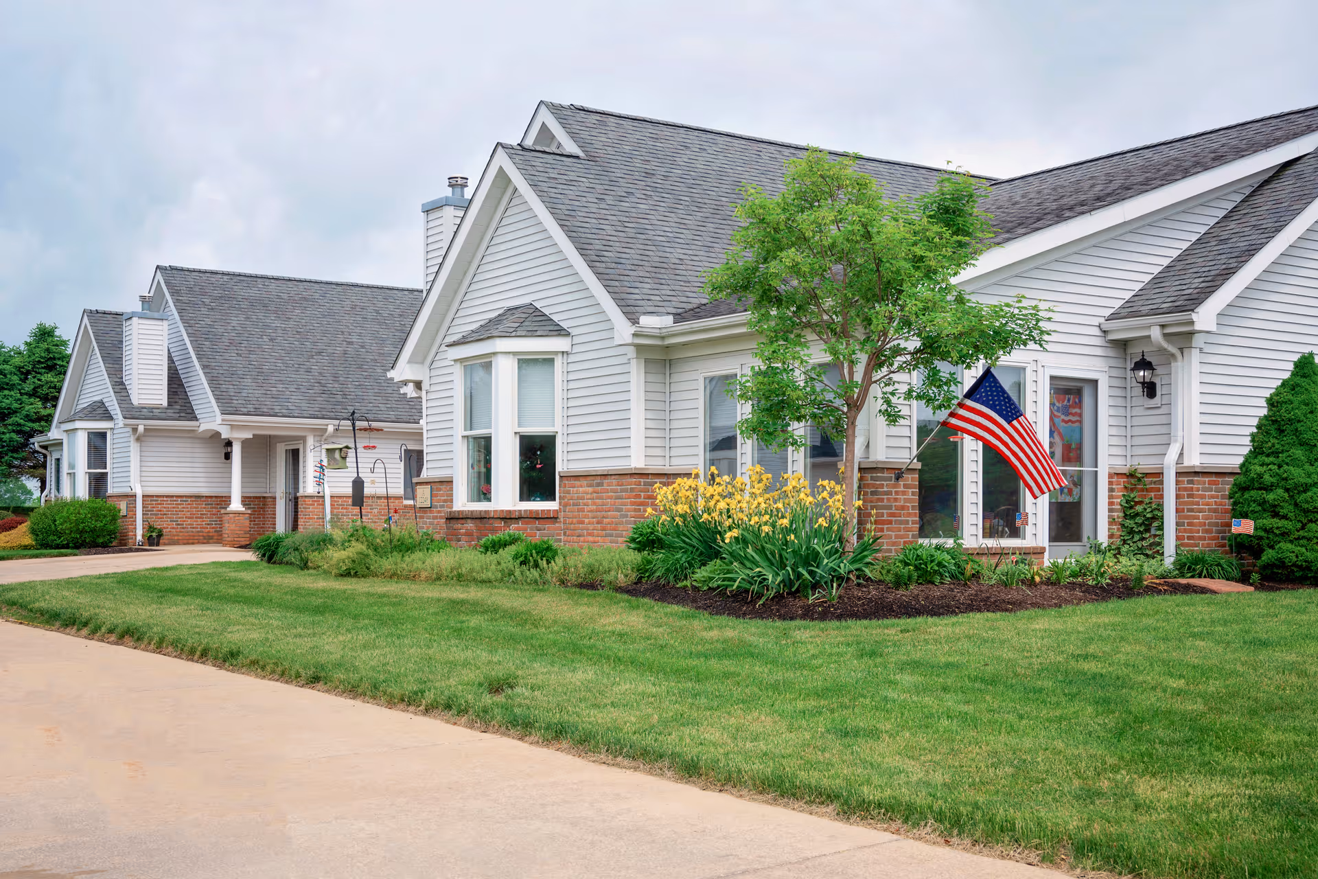 Exterior view of a residential-style building with white siding and brick accents, featuring a well-maintained lawn, a small tree, yellow flowers, and an American flag mounted near the entrance.