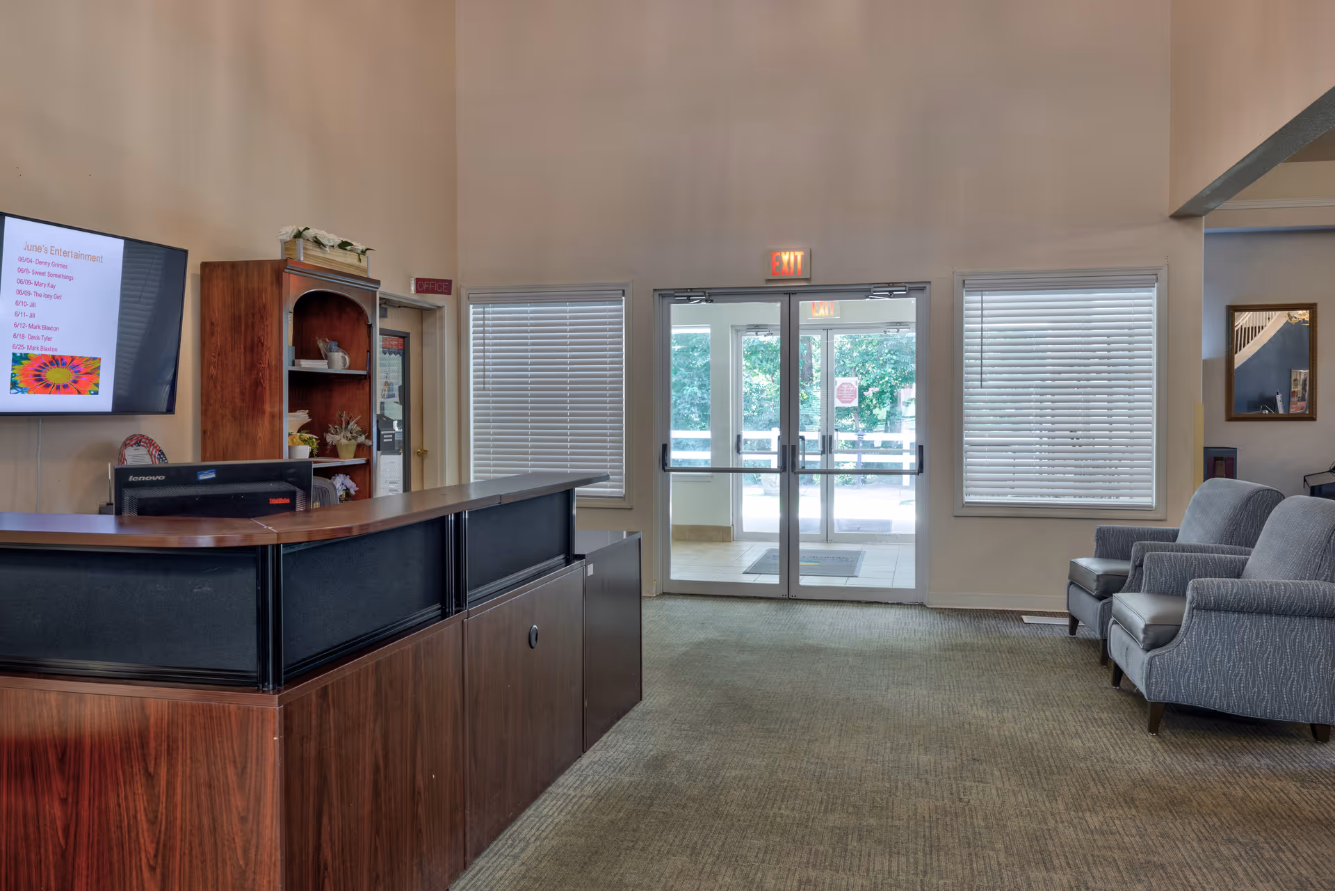 Reception area of TerraBella Riverplace with a wooden front desk, a computer monitor, a wall-mounted TV displaying a schedule, a wooden shelf with decorative items, two gray armchairs, windows with blinds, and glass double doors leading outside.