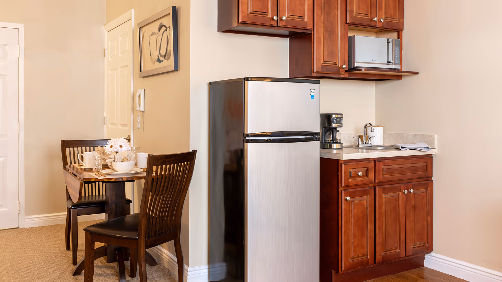 A small kitchen area with wooden cabinets, a stainless steel refrigerator, a coffee maker, and a sink. Adjacent to the kitchen is a dining table set for two with dark wooden chairs and a floral centerpiece. The walls are light-colored, and there is a framed abstract artwork hanging above the table.