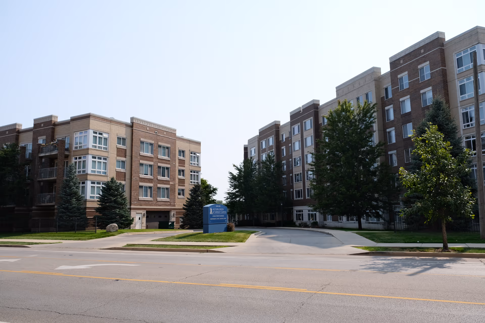 Exterior view of Senior Suites of Autumn Green at Wright Campus, showing two multi-story brick buildings with numerous windows, trees, and a driveway entrance with a blue sign in the center.