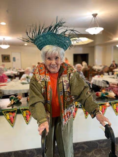 An elderly woman wearing a colorful jacket and a green feathered hat is smiling and using a walker in a dining room decorated with festive banners and maracas. Other elderly people are seated at tables in the background.