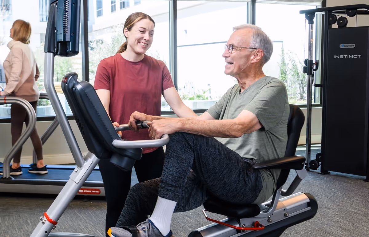 An elderly man exercising on a seated rowing machine in a fitness center, assisted by a smiling female trainer. Another person is walking on a treadmill in the background near large windows.