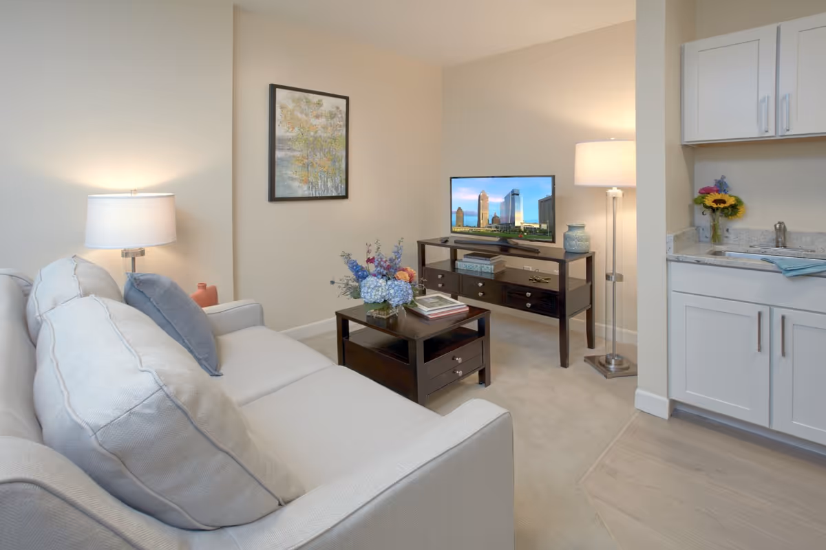A cozy living room area in a senior living facility featuring a light gray sofa with blue and beige cushions, a dark wood coffee table with a flower arrangement and books, a matching TV stand with a flat-screen TV displaying a cityscape, a floor lamp, a table lamp, a framed painting on the wall, and a small kitchenette area with white cabinets and a granite countertop with a vase of flowers.