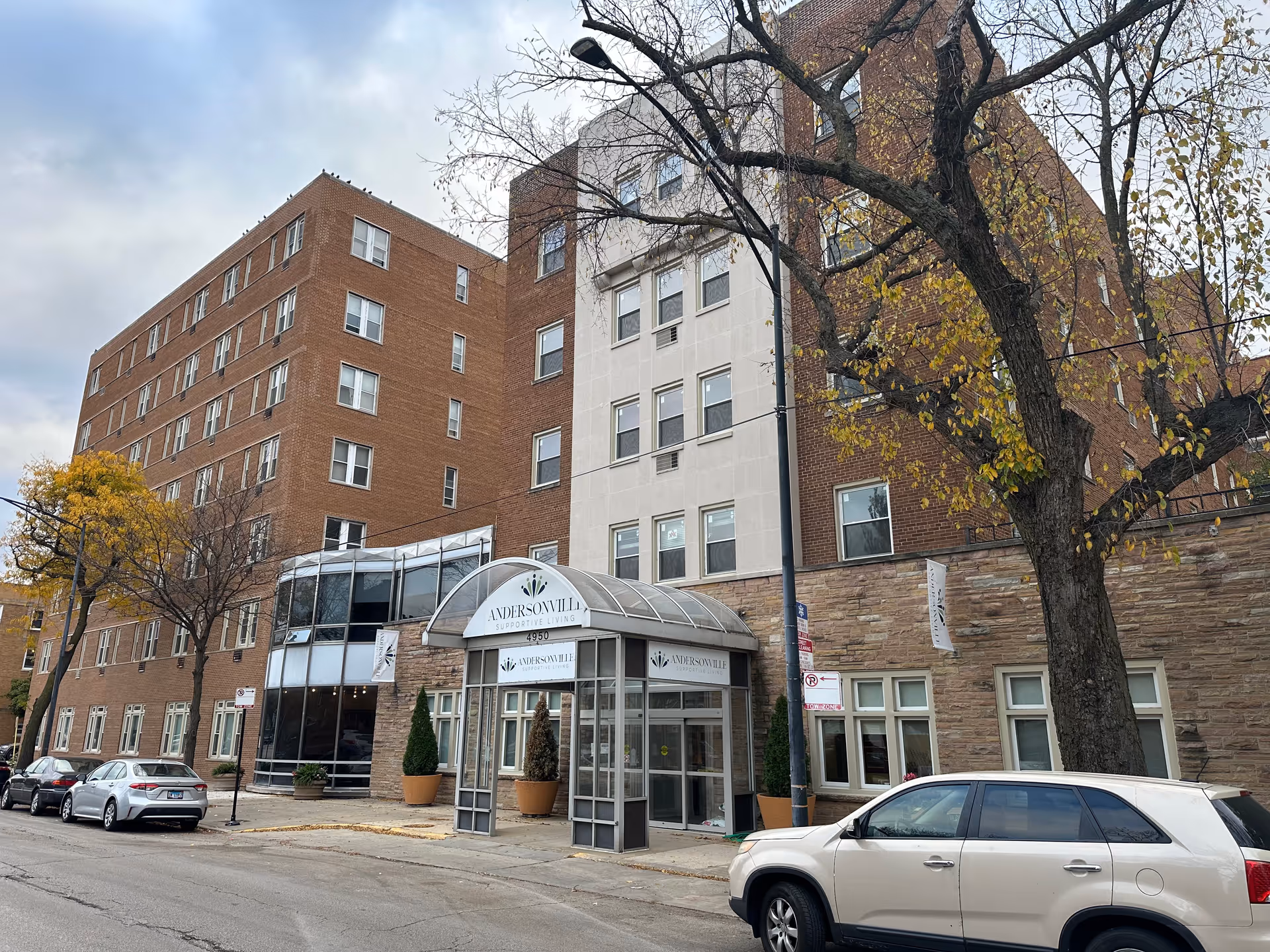 Multi-story brick retirement community building with a glass-covered entrance and cars parked along the street.
