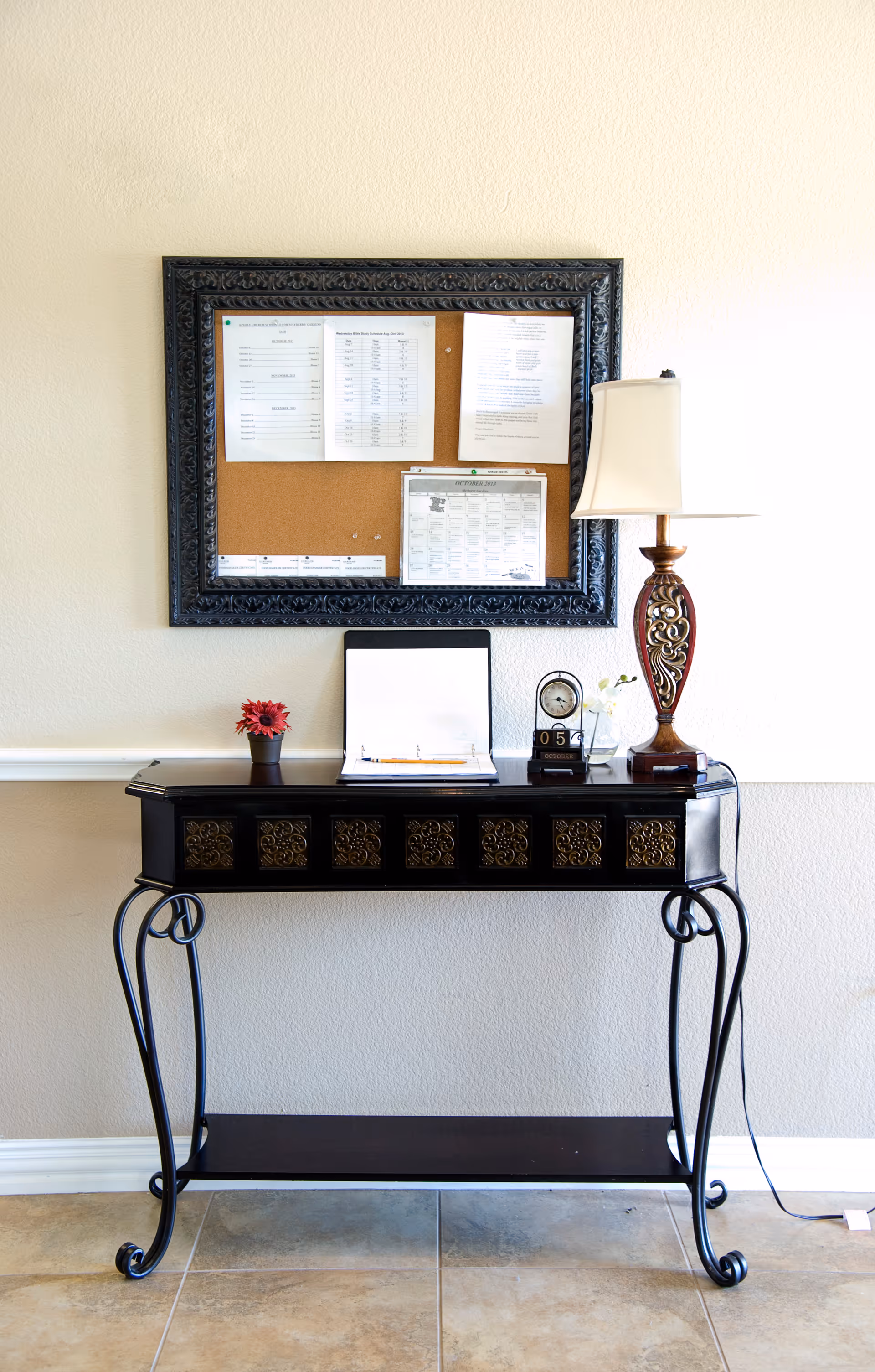 A decorative black console table with ornate gold panels and curved legs stands against a beige wall. On the table are a small potted red flower, an open binder with papers, a vintage-style clock, a small glass vase with a white flower, and a decorative table lamp with a white shade. Above the table is a large framed cork bulletin board with several pinned papers and a calendar.