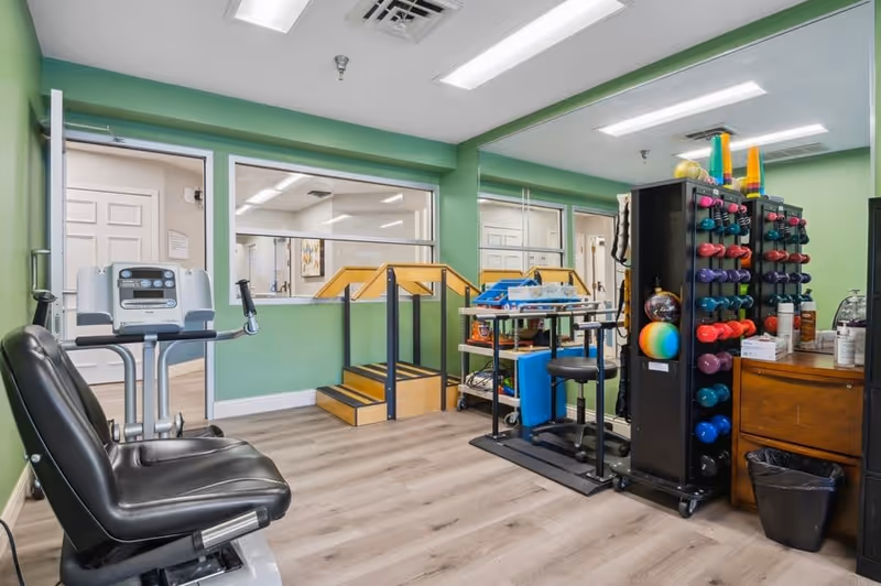 A small exercise room with light green walls and wood flooring. The room contains various exercise equipment including a stationary bike, a rack of colorful dumbbells, therapy balls, and a small set of wooden stairs with handrails. There is a large mirror on one wall and windows looking into an adjacent room.
