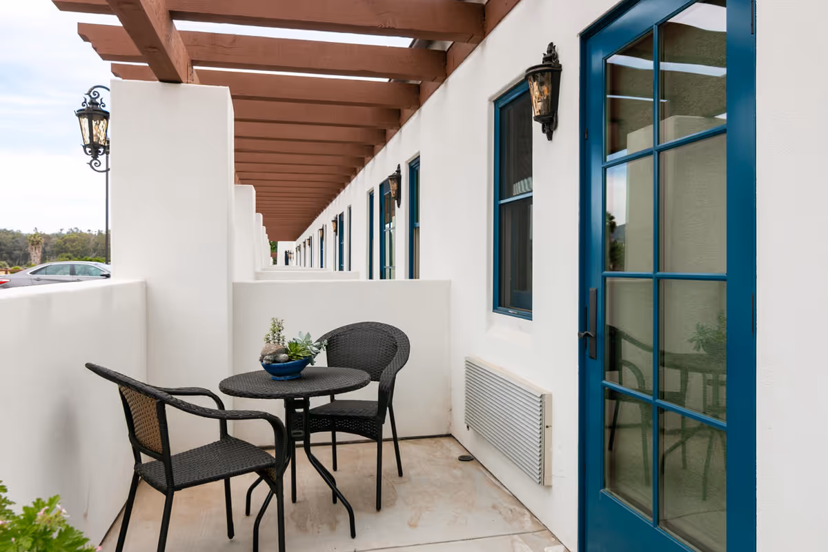 Small private patio with a round table and two wicker chairs beside a blue-framed glass door and windows under a wooden pergola.