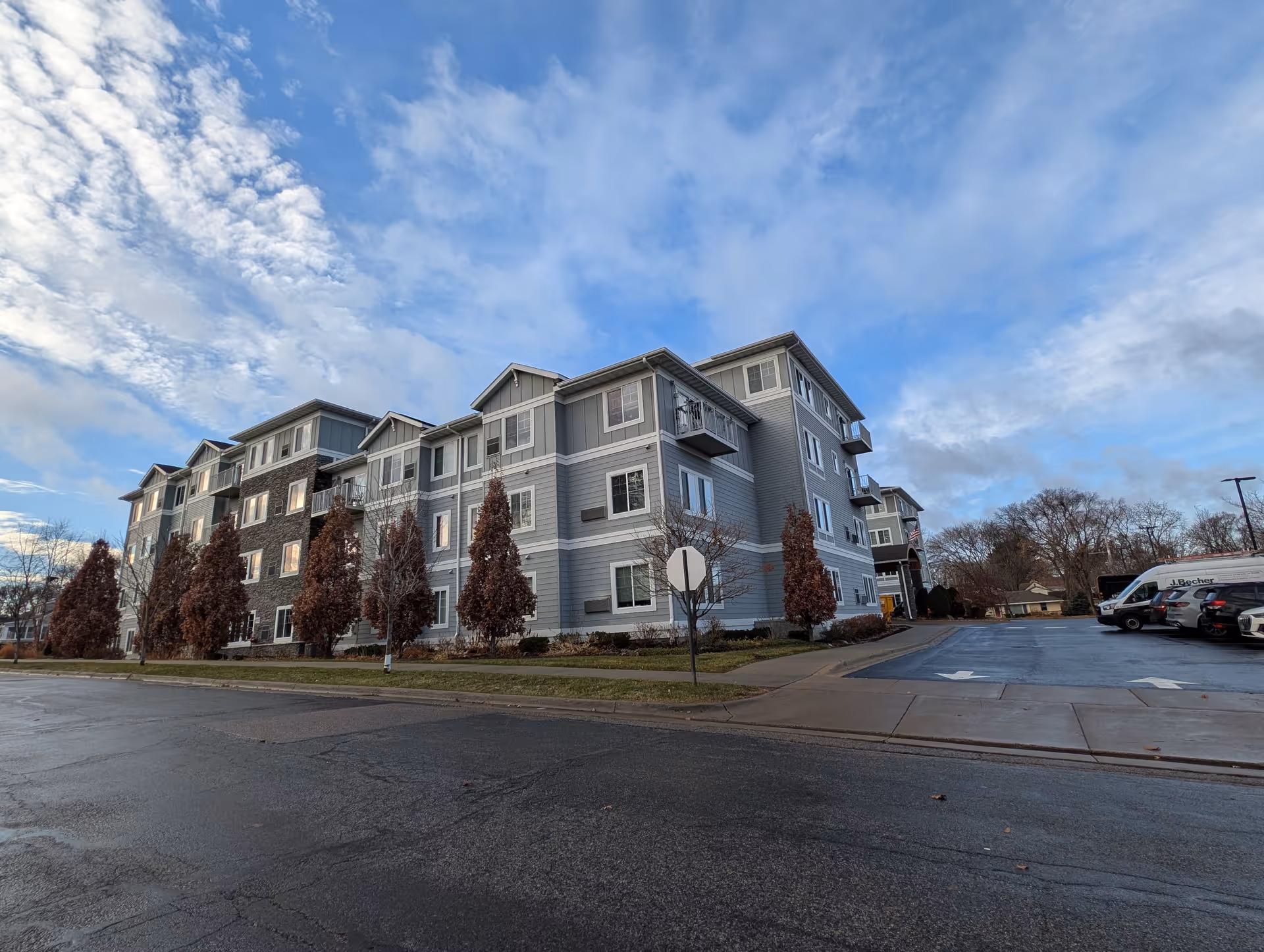 A four-story gray and stone-clad senior living building with trimmed trees and a parking lot under a partly cloudy blue sky.