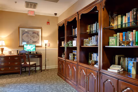 A cozy interior room featuring a large wooden bookshelf filled with books and decorative items on the right side. On the left side, there is a wooden desk with a computer monitor, a chair, a lamp, and a framed picture on the wall above the desk. The room has carpeted flooring and warm lighting.