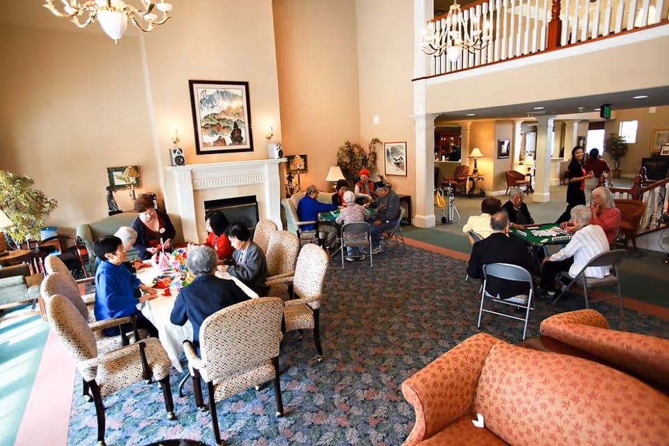 A spacious living room area in a senior living facility with multiple groups of elderly people sitting around tables engaged in activities and games. The room features comfortable upholstered chairs, a fireplace with artwork above it, and warm lighting. There is a balcony railing visible on the upper level and a woman standing near the back of the room.