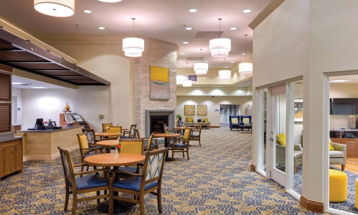 A spacious and well-lit common area in a senior living facility featuring round wooden tables with chairs, a fireplace with a modern painting above it, patterned carpet, and multiple pendant lights hanging from the ceiling. There is a counter area to the left and a glass-walled room with seating to the right.