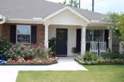 Front exterior view of a single-story brick house with a black front door, white porch columns, a small porch with a white rocking chair, and well-maintained flower beds and lawn.