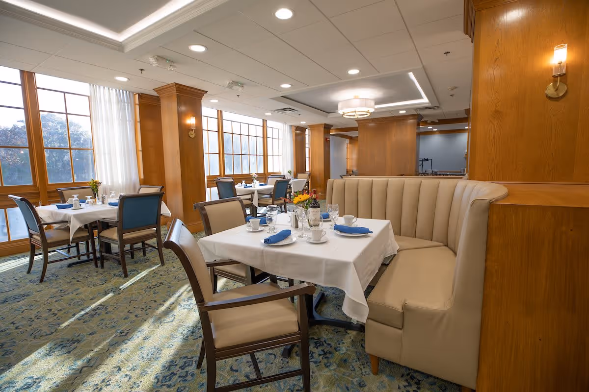 A bright dining room with large windows letting in natural light. The room features tables covered with white tablecloths, set with cups, glasses, napkins, and a bottle of wine. There are upholstered chairs and a beige curved booth seating area. The walls and columns are wood-paneled, and the ceiling has recessed lighting and a modern chandelier.