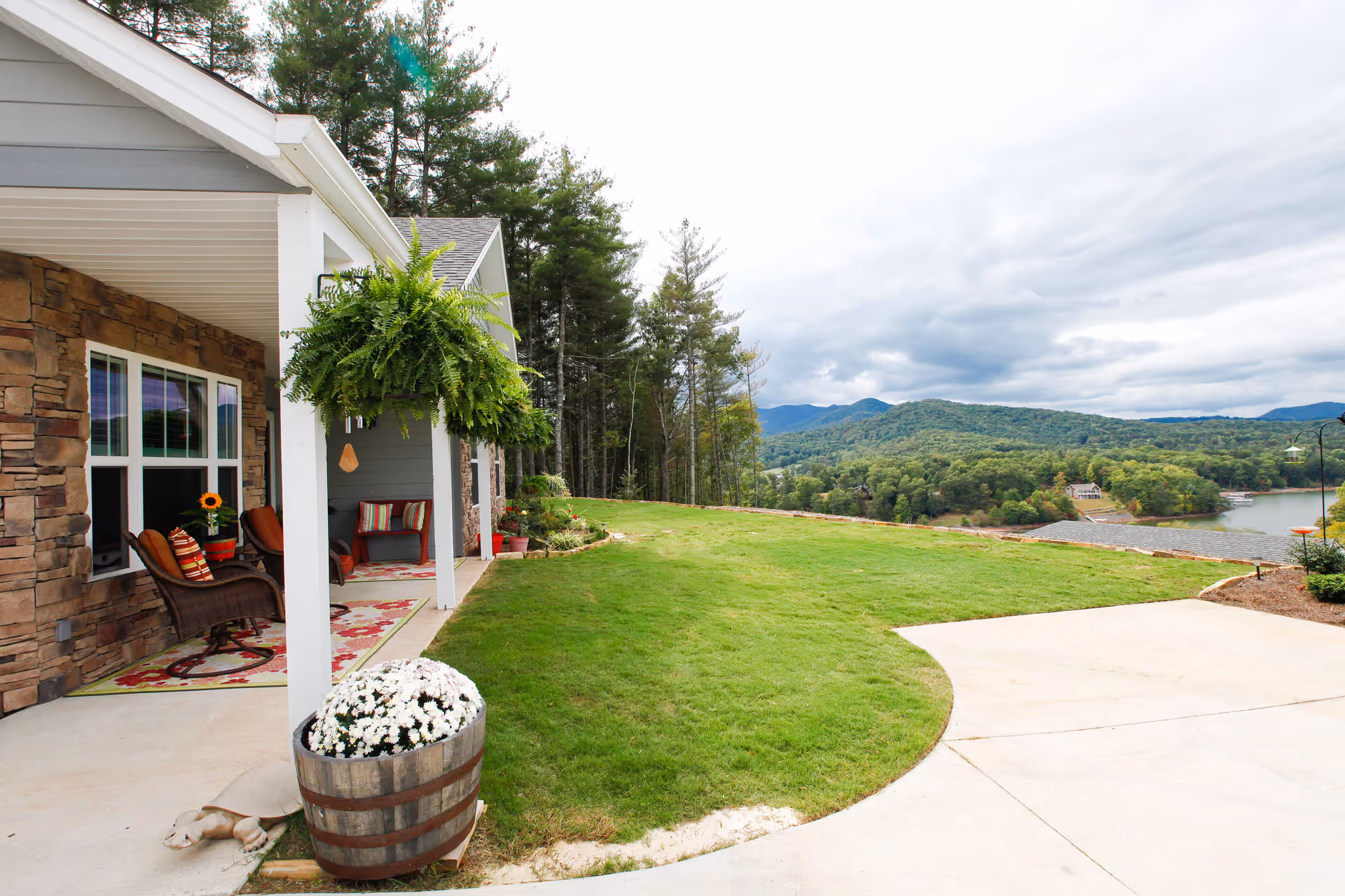 A porch area of a building with stone and gray siding, featuring wicker chairs with cushions, a hanging fern plant, and a wooden barrel planter with white flowers. The porch overlooks a grassy lawn with trees and hills in the background under a cloudy sky.