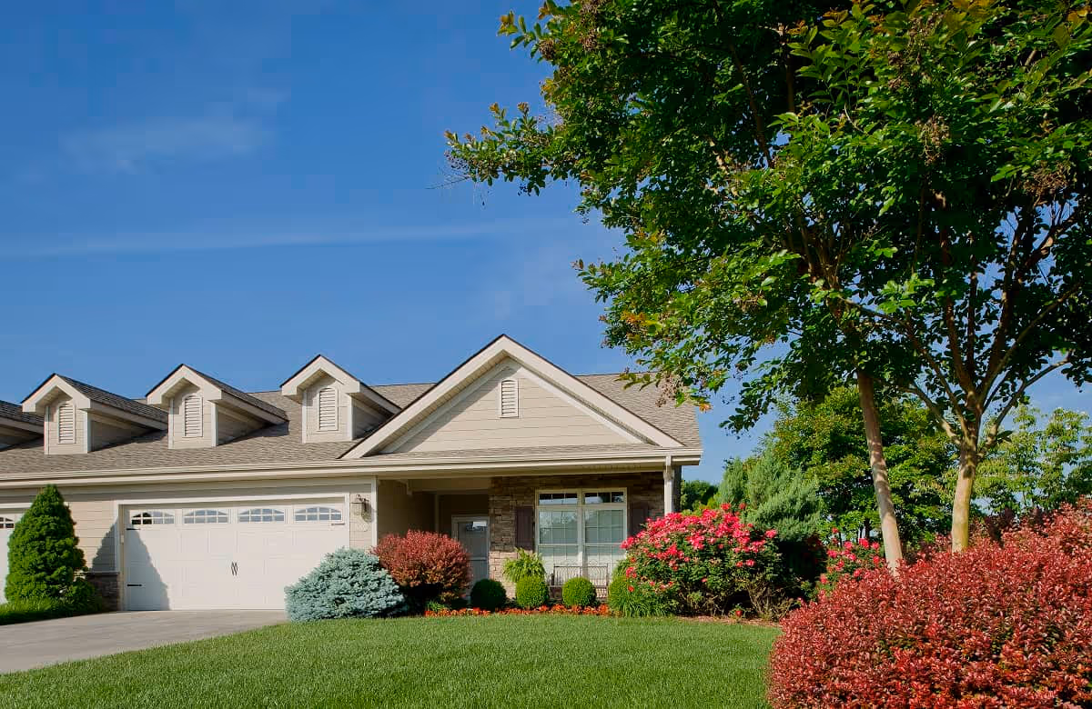 Exterior view of a single-story residential building with a garage, surrounded by a well-maintained lawn, colorful bushes, and trees under a clear blue sky.