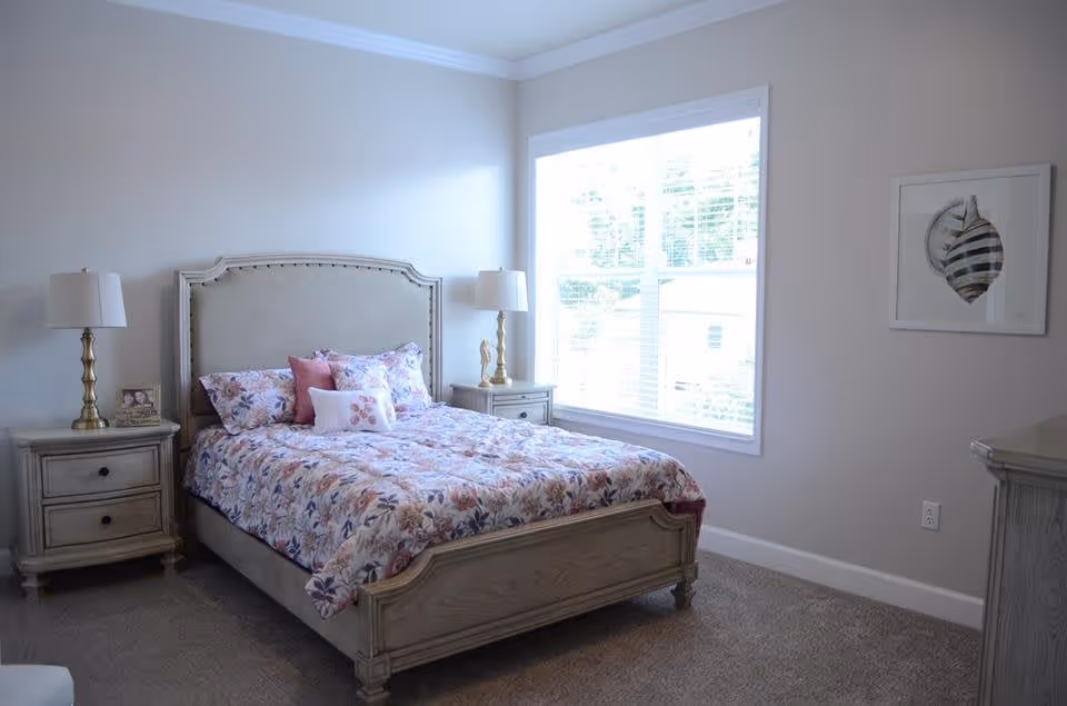 A bright bedroom with a large window letting in natural light. The room features a wooden bed with a floral patterned comforter and several pillows. There are matching wooden nightstands on either side of the bed, each with a lamp. A framed seashell artwork hangs on the wall to the right.