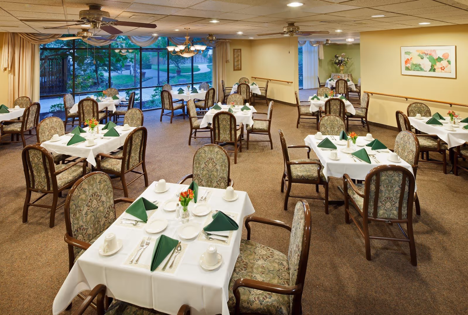 Dining room with multiple tables set with white tablecloths, green napkins, and floral centerpieces overlooking a garden through large windows.