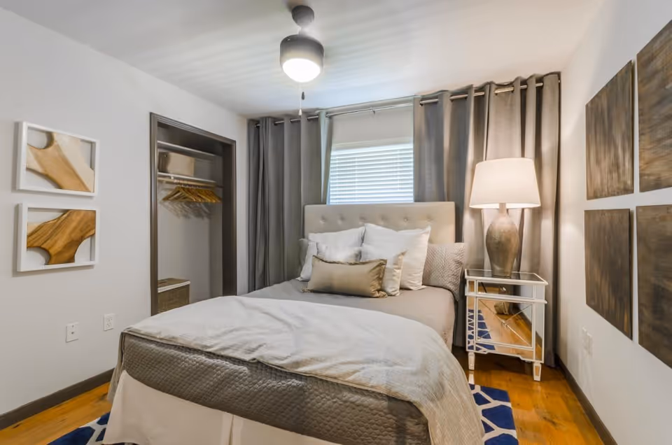 Small furnished bedroom with a bed, pillows, bedside lamp on a mirrored nightstand, open closet and curtains over a window.
