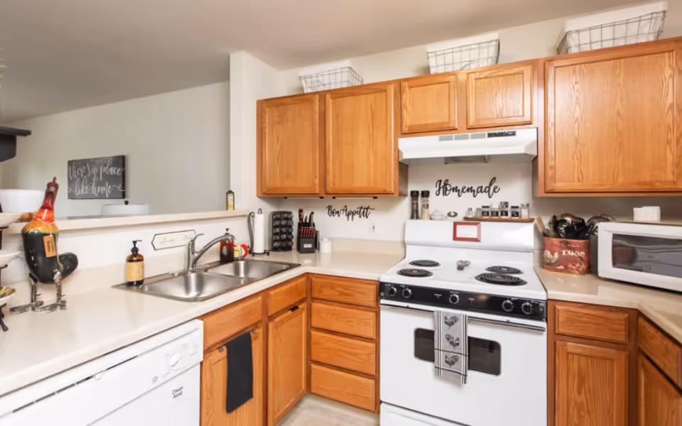 A compact kitchen with wooden cabinets, a white electric stove, double sink, microwave and various countertop items.