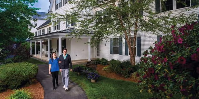 Two people walk along a landscaped path in front of a white multi-story senior living building with a porch.