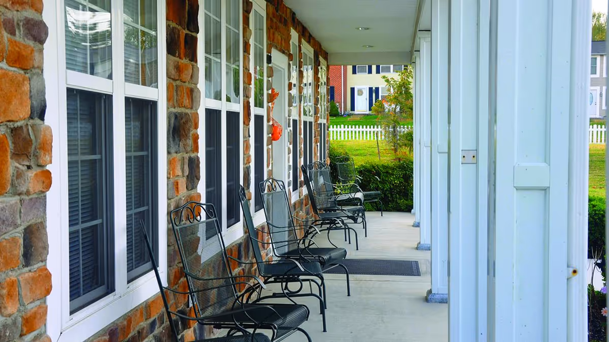 A covered outdoor porch area with several black metal chairs lined up against a stone wall with multiple windows. The porch has white pillars and overlooks a green lawn with a white picket fence and houses in the background.